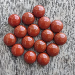 Può includere: Una collezione di sedici pietre preziose rotonde e lucide di diaspro rosso. Le pietre sono disposte su una superficie di legno grigio usurato. Ogni pietra ha una superficie liscia e riflettente.