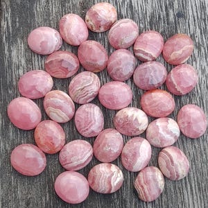 May include: A collection of oval-shaped, polished rhodochrosite stones. The stones display a range of pink hues, from pale to deep rose, with distinctive banding patterns. The gemstones are arranged on a weathered wooden surface.