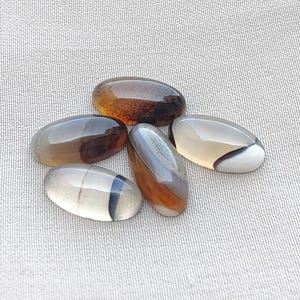 Könnte beinhalten: Fünf glatte, oval geformte Cabochons mit polierten Oberflächen. Die Steine weisen durchscheinende und undurchsichtige Bereiche in Bernstein-, Braun-, Grau- und Weißtönen auf und zeigen natürliche interne Bänderungen, Farbverläufe und dunkle Einschlüsse.