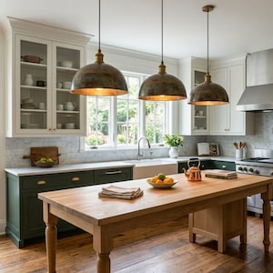 Pode incluir: Uma cozinha bem iluminada com uma ilha de madeira e armários verde escuro. Três luminárias pendentes de bronze pendem sobre a ilha. Uma janela fornece luz natural. A cozinha apresenta uma pia de fazenda e eletrodomésticos de aço inoxidável.