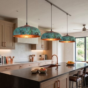 Puede incluir: Tres lámparas colgantes en forma de cúpula suspendidas de un sistema de rieles. Cada luminaria presenta un exterior de cobre oxidado texturizado con un acabado de pátina verde y azul, que contrasta con una superficie interior metálica pulida.