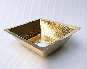 May include: A square-shaped sink with a polished gold-tone finish and a distinct hammered texture across its interior surfaces. The basin features a central circular drain opening and wide, flat edges along the top rim for installation.