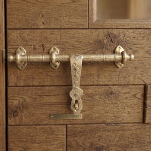 Op de afbeelding: Een messing deurgrendel met een ingewikkeld gegraveerd patroon gemonteerd op een donkerhouten deur. De horizontale schuifbalk heeft decoratieve banden en de verticale sluitplaat toont een geschulpt ontwerp. Een klein rechthoekig plaatje vermeldt Artisan Hardware.