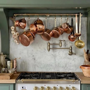 Puede incluir: Una cocina con ollas y sartenes de cobre colgadas de una barra de latón. La imagen muestra una estufa blanca con detalles de latón, una placa para salpicaduras con efecto mármol y gabinetes verdes. Un manojo de ajo cuelga a la izquierda.