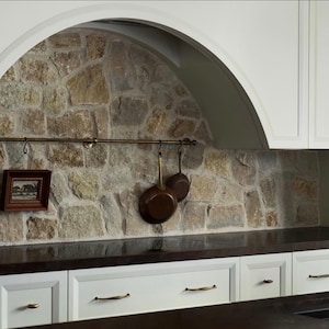 May include: A kitchen with white cabinets, brass handles, and a dark work surface. A stone wall forms the backsplash, with a brass rail holding copper pans and a framed picture. The design has a rustic, farmhouse aesthetic.