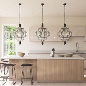 May include: Three black geometric pendant lights suspended over a kitchen island. The lights have a chain and a multi-faceted glass design. The kitchen features a light wood island with a white countertop, stools, and a window.