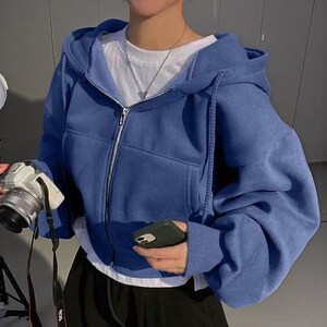 Op de afbeelding: Een blauwe hoodie met ritssluiting, capuchon met trekkoord en voorzakken. De hoodie wordt gedragen over een wit t-shirt. Een zilveren ketting is zichtbaar. De persoon houdt een camera en een telefoon vast.