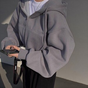 Op de afbeelding: Een grijze hoodie met ritssluiting, trekkoord en een voorzak. De hoodie wordt gedragen met een wit shirt en een zwarte broek. Een zwarte telefoon wordt vastgehouden en een camerariem is zichtbaar.
