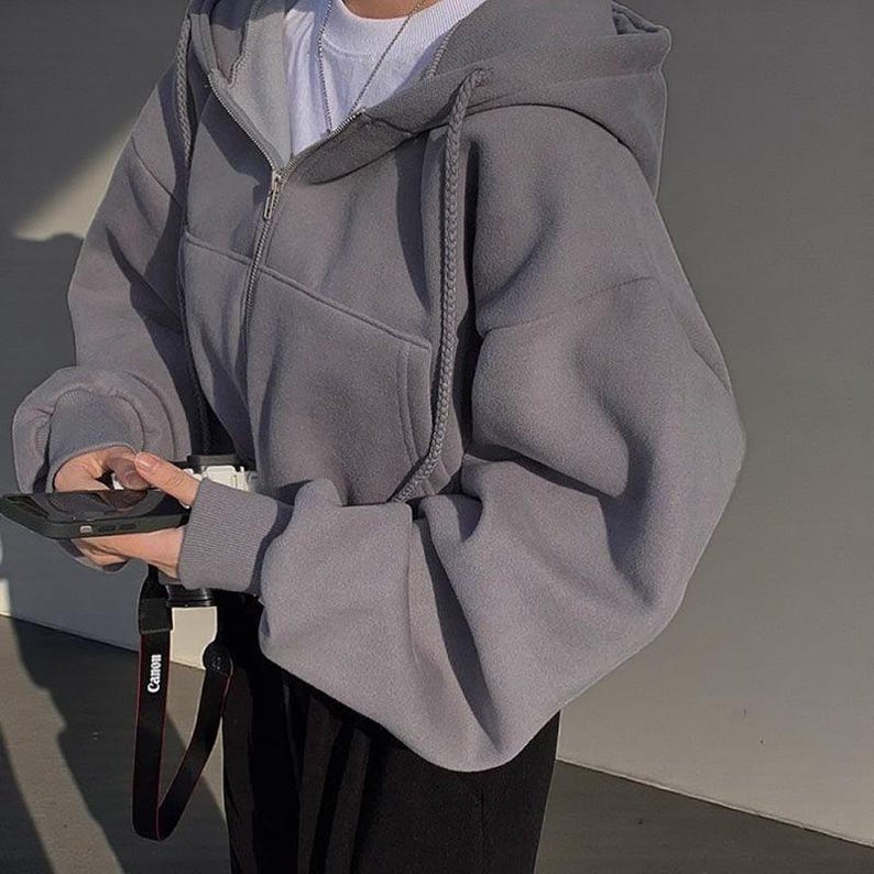 Op de afbeelding: Een grijze hoodie met ritssluiting, trekkoord en een voorzak. De hoodie wordt gedragen met een wit shirt en een zwarte broek. Een zwarte telefoon wordt vastgehouden en een camerariem is zichtbaar.