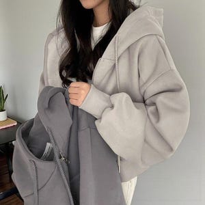 Op de afbeelding: Een lichtgrijze hoodie met ritssluiting, capuchon met trekkoord en geribde manchetten. De hoodie wordt door een persoon omhooggehouden. Een tweede, donkerdere grijze hoodie wordt in de andere hand van de persoon gehouden. De achtergrond is een effen, lichtgrijze muur.