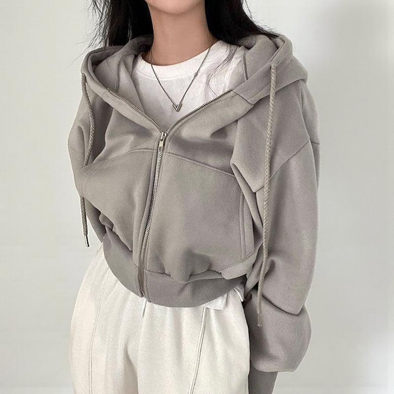 Op de afbeelding: Een lichtgrijze hoodie met ritssluiting, capuchon met trekkoord en voorzakken. De hoodie wordt gedragen over een wit T-shirt en gecombineerd met een witte broek. De rits is zilverkleurig. De algehele stijl is casual en comfortabel.