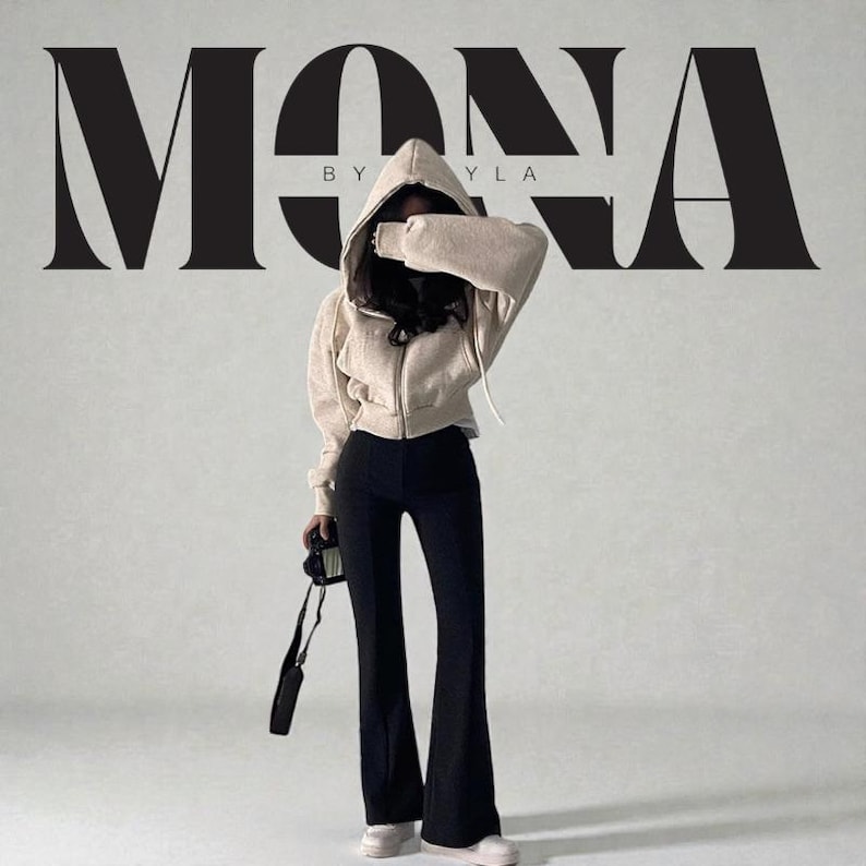 Op de afbeelding: Een vrouw draagt een beige hoodie met rits, een zwarte flared broek en witte sneakers. De afbeelding toont de tekst "MONA BY YLA" in een vetgedrukte, gestileerde lettertype. Een camera wordt in de hand van de vrouw gehouden.