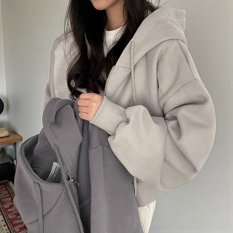 Op de afbeelding: Een lichtgrijze hoodie met ritssluiting, een capuchon met trekkoord en een bijpassende grijze hoodie. De hoodies zijn gemaakt van een zacht, comfortabel materiaal. De hoodie met ritssluiting heeft een volledige ritssluiting en geribde manchetten.