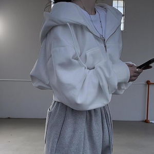 Op de afbeelding: Een witte hoodie met ritssluiting, capuchon met trekkoord en voorzakken. De hoodie wordt gecombineerd met een grijze joggingbroek. De outfit is casual en comfortabel, geschikt voor dagelijks gebruik.