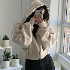 Op de afbeelding: Een beige hoodie met een volledige ritssluiting. De hoodie heeft een capuchon met trekkoord en lange mouwen. De persoon op de foto draagt een zwarte broek. De achtergrond is een witte muur en een raam.