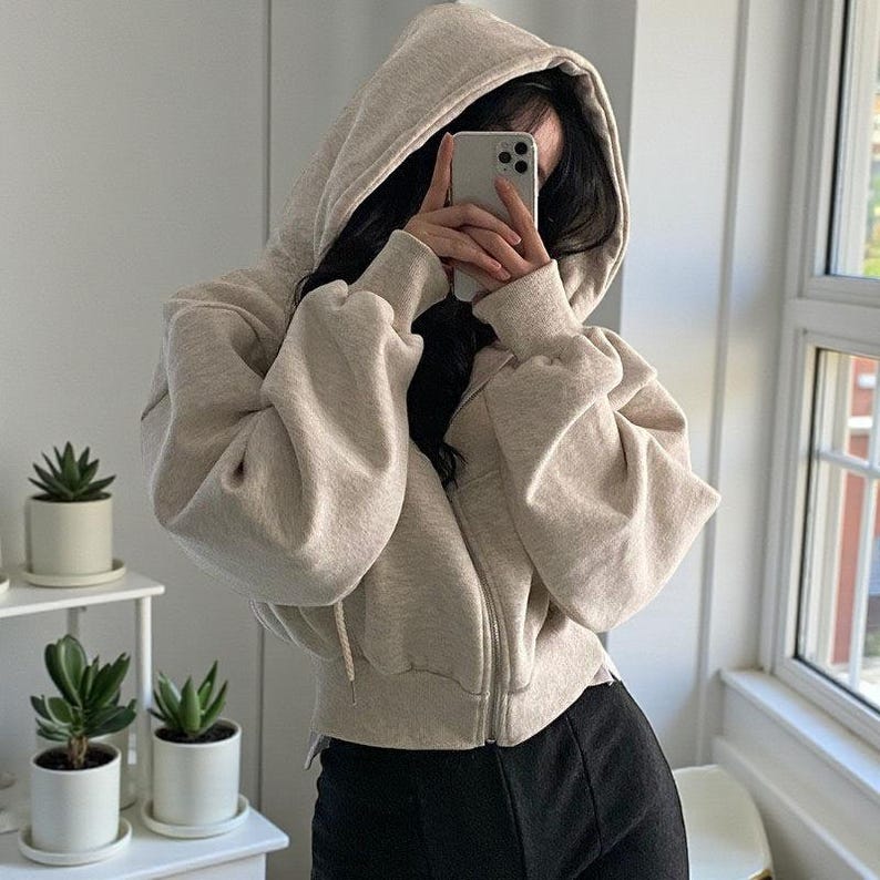 Op de afbeelding: Een beige hoodie met een volledige ritssluiting. De hoodie heeft een capuchon met trekkoord en lange mouwen. De persoon op de foto draagt een zwarte broek. De achtergrond is een witte muur en een raam.