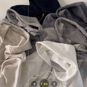 Op de afbeelding: Een verzameling hoodies met ritssluiting in verschillende neutrale kleuren, waaronder wit, grijs en marineblauw. De hoodies zijn gemaakt van een zacht materiaal en hebben trekkoorden en voorzakken. De afbeelding is van bovenaf genomen en toont de textuur en het ontwerp van de kleding.