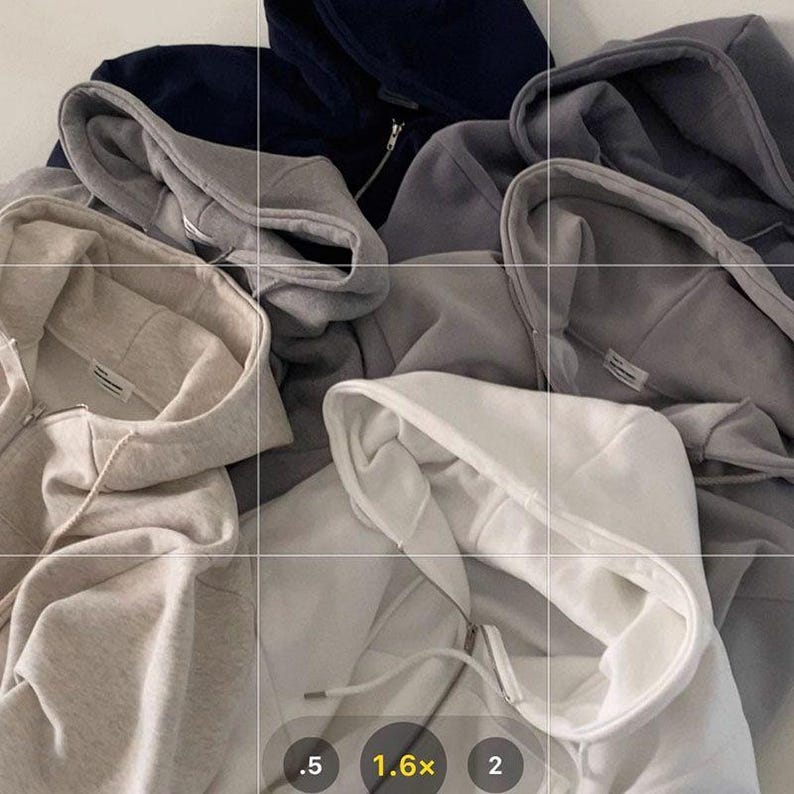 Op de afbeelding: Een verzameling hoodies met ritssluiting in verschillende neutrale kleuren, waaronder wit, grijs en marineblauw. De hoodies zijn gemaakt van een zacht materiaal en hebben trekkoorden en voorzakken. De afbeelding is van bovenaf genomen en toont de textuur en het ontwerp van de kleding.