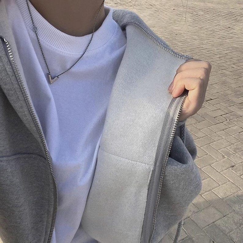 Op de afbeelding: Een grijze hoodie met ritssluiting, witte binnenkant en zilveren rits. De hoodie wordt gedragen over een wit t-shirt en een zilveren ketting. De afbeelding is een close-up, die de textuur van de stof en de details van de rits laat zien.