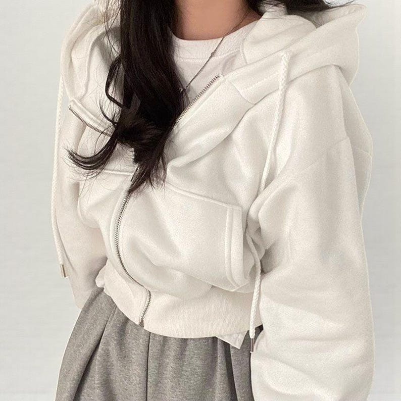 Op de afbeelding: Een witte hoodie met ritssluiting, capuchon met trekkoord en voorzakken. De hoodie wordt gecombineerd met een grijze joggingbroek en een wit t-shirt eronder. De rits is zilverkleurig.