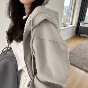 Op de afbeelding: Een lichtgrijze hoodie met ritssluiting en capuchon met trekkoord. De hoodie heeft een ritssluiting over de hele lengte en is gemaakt van een zacht, comfortabel materiaal. De hoodie wordt gecombineerd met een wit t-shirt.