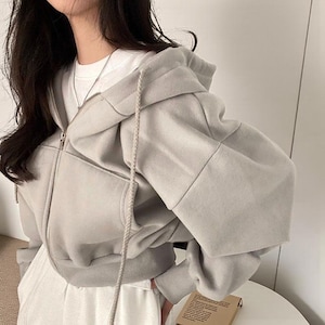 Op de afbeelding: Een lichtgrijze hoodie met ritssluiting, capuchon met trekkoord en voorzakken. De hoodie heeft een cropped design en lange mouwen. De rits is zilverkleurig. De persoon draagt er een wit t-shirt onder.
