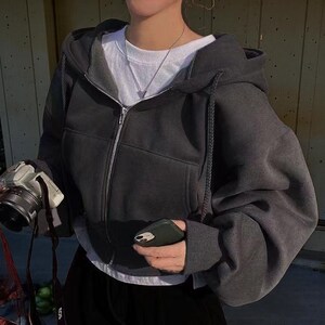Op de afbeelding: Een donkergrijze hoodie met ritssluiting, capuchon met trekkoord en voorzakken. De hoodie wordt gedragen over een wit t-shirt. Een zilveren rits loopt over de voorkant. De persoon houdt een camera en een telefoon vast.