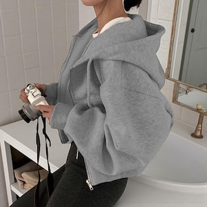 Op de afbeelding: Een lichtgrijze, korte hoodie met ritssluiting en een grote capuchon. De hoodie heeft een volledige ritssluiting en lange mouwen. Het model draagt er een wit shirt onder en een donkergrijze broek. Een camera wordt in de handen van het model gehouden.