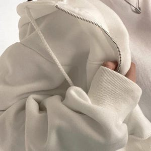 Op de afbeelding: Een close-up van een witte hoodie met een rits en trekkoord. De stof van de hoodie is zacht en getextureerd, met geribde manchetten en een capuchon. De rits is zilverkleurig en het trekkoord is wit.