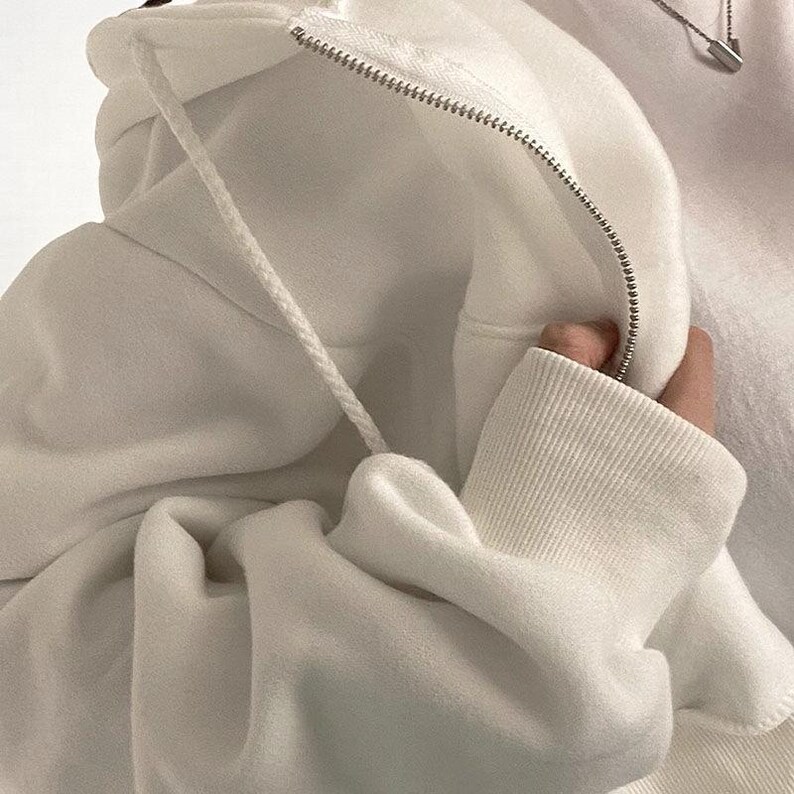 Op de afbeelding: Een close-up van een witte hoodie met een rits en trekkoord. De stof van de hoodie is zacht en getextureerd, met geribde manchetten en een capuchon. De rits is zilverkleurig en het trekkoord is wit.