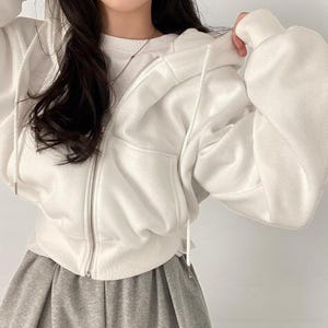 Op de afbeelding: Een witte hoodie met ritssluiting, capuchon met trekkoord en voorzakken. De hoodie wordt gecombineerd met een grijze rok. De rits is zilverkleurig. De mouwen zijn lang en het materiaal lijkt zacht te zijn.