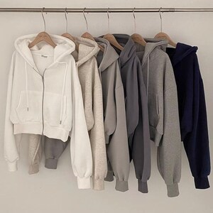 Op de afbeelding: Zes hoodies met ritssluiting in diverse neutrale kleuren, waaronder wit, beige, grijs en marineblauw, hangen aan een metalen stang. Elke hoodie heeft een capuchon, voorzakken en geribde manchetten. De hoodies zijn gemaakt van een zachte stof.