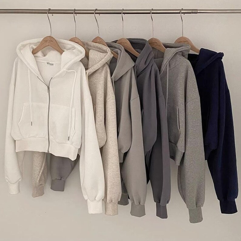 Op de afbeelding: Zes hoodies met ritssluiting in diverse neutrale kleuren, waaronder wit, beige, grijs en marineblauw, hangen aan een metalen stang. Elke hoodie heeft een capuchon, voorzakken en geribde manchetten. De hoodies zijn gemaakt van een zachte stof.