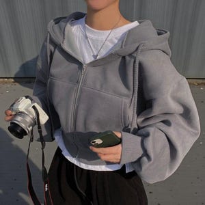 Op de afbeelding: Een grijze hoodie met ritssluiting, capuchon met trekkoord en voorzakken. De hoodie wordt gedragen over een wit t-shirt. Een camera met een zwarte band en een groene telefoon zijn ook zichtbaar.