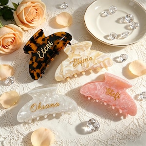May include: Four hair claw clips in tortoise shell, cream, white, and pink marble patterns. Each clip features a gold-toned name engraved on the surface: Eleni, Elyssia, Oksana, and Stefi. The clips have a curved design with multiple teeth.