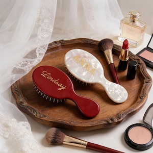 May include: Two oval hairbrushes with gold lettering, one in a glossy deep red with the name Lindsay and one in a pearlescent white finish with the name Anna, accompanied by makeup brushes, a lipstick, and a compact on a wooden tray.