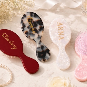 May include: Four oval hairbrushes with handles. One is solid dark red with the name Lindsay in gold. One is black and white tortoiseshell with the name Brenna in gold. One is white pearlescent with the name Anna in gold. One is light pink pearlescent.