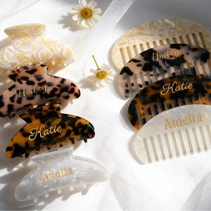 May include: A collection of four hair claw clips and four hair combs made of marbled and tortoiseshell resin. The accessories feature gold-coloured personalised names including Hailey, Katie, and Amelia printed on the surface of each item.