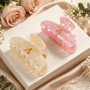 May include: Two hair claw clips with a marbled finish, one in cream and one in translucent pink. Each clip features a gold-colored letter A centered on the top surface. The clips have a curved, multi-pronged design for securing hair in place.