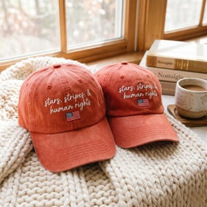 May include: Two terracotta-colored baseball caps with the embroidered text "stars, stripes, & human rights" and a small American flag patch. The caps are displayed on a cream-colored knitted blanket near a window.