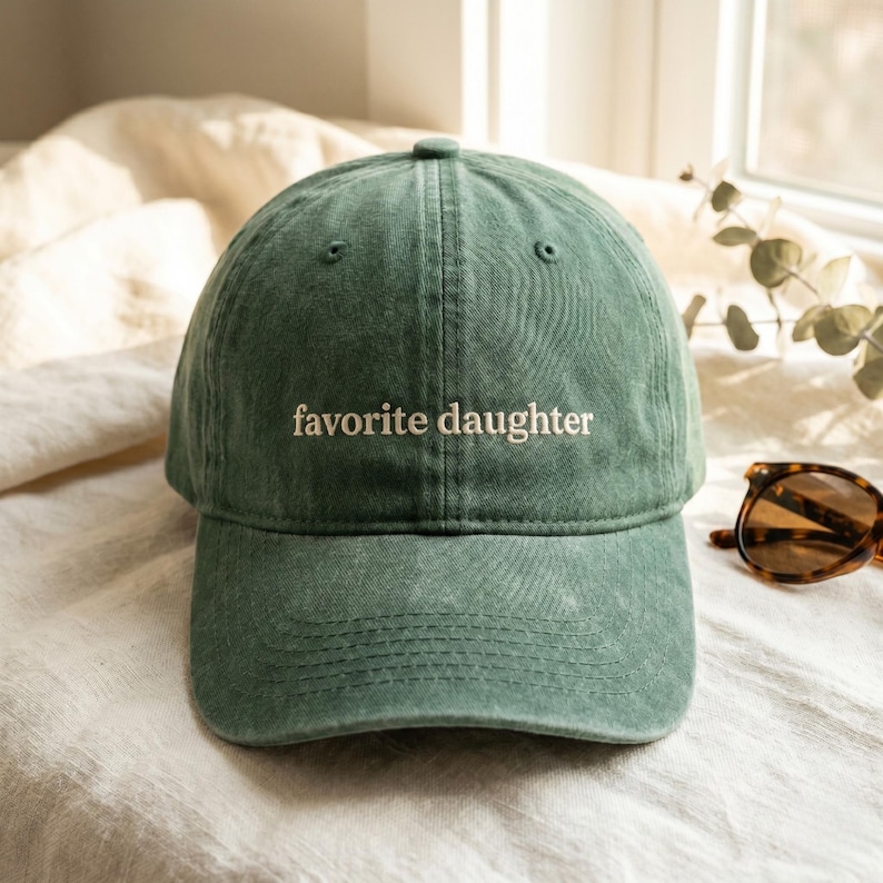 K&ouml;nnte beinhalten: Eine gr&uuml;ne Baseballkappe mit dem gestickten Text "favorite daughter" in Wei&szlig;. Die Kappe ist aus einem weichen, strukturierten Stoff gefertigt und hat einen gebogenen Schirm. Im Hintergrund ist eine Sonnenbrille zu sehen.