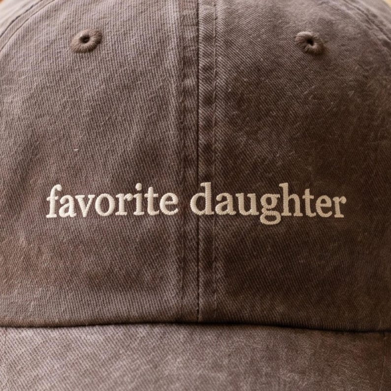 May include: A brown baseball cap with the embroidered text "favorite daughter" in white. The cap has a curved brim and a fabric strap for adjustment. The cap is made of a soft, durable material.