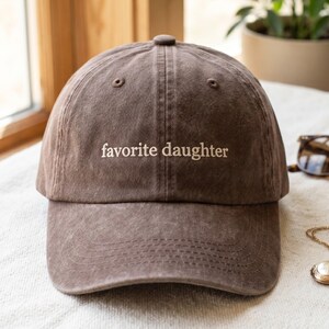 May include: A brown baseball cap with the embroidered text "favorite daughter" in white. The cap has a curved brim and a classic six-panel design. The hat is made of a soft, worn material.