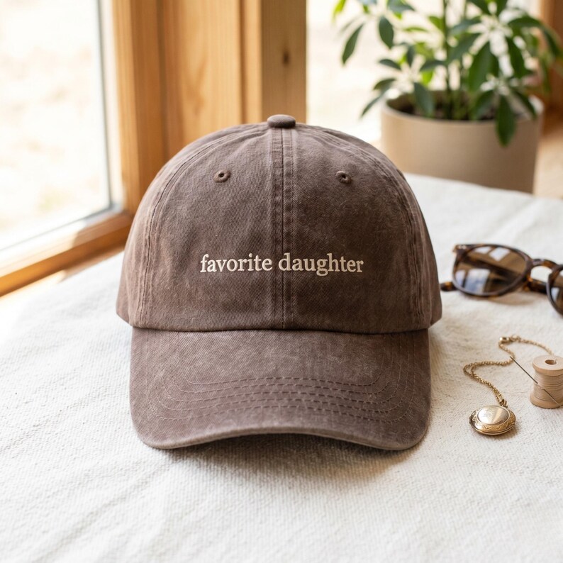 May include: A brown baseball cap with the phrase "favorite daughter" embroidered in white. The cap is made of a soft, textured material and has a curved brim. The cap is sitting on a white surface.