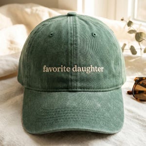 K&ouml;nnte beinhalten: Gr&uuml;ne Baseballkappe mit dem gestickten Text "favorite daughter" in Wei&szlig;. Die Kappe hat einen gebogenen Schirm und einen Stoffriemenverschluss. Der Hut ist aus einem weichen, strukturierten Material gefertigt.