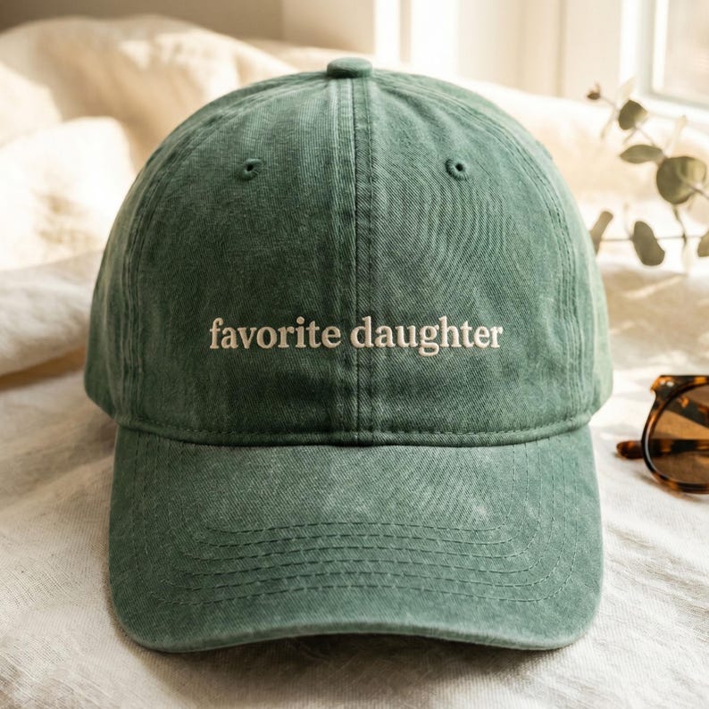 K&ouml;nnte beinhalten: Gr&uuml;ne Baseballkappe mit dem gestickten Text "favorite daughter" in Wei&szlig;. Die Kappe hat einen gebogenen Schirm und einen Stoffriemenverschluss. Der Hut ist aus einem weichen, strukturierten Material gefertigt.