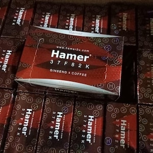 May include: Close-up of multiple boxes of Hamer ginseng and coffee. The boxes are primarily brown and red with the brand name and product information printed in white. The boxes are arranged in a grid pattern.