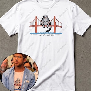 Puede incluir: Una camiseta blanca de manga corta hecha de 100% algodón con un estampado gráfico de un gato atigrado gris sentado en el puente Golden Gate con el texto SAN FRANCISCO impreso debajo. La camiseta tiene cuello redondo y corte estándar.