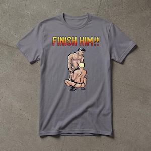 May include: A grey short-sleeved t-shirt featuring a pixel art graphic of two figures in a wrestling pose. Above the graphic, the text FINISH HIM!! is printed in a yellow and red gradient font. The shirt has a crew neckline and a standard fit.