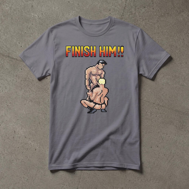 May include: A grey short-sleeved t-shirt featuring a pixel art graphic of two figures in a wrestling pose. Above the graphic, the text FINISH HIM!! is printed in a yellow and red gradient font. The shirt has a crew neckline and a standard fit.